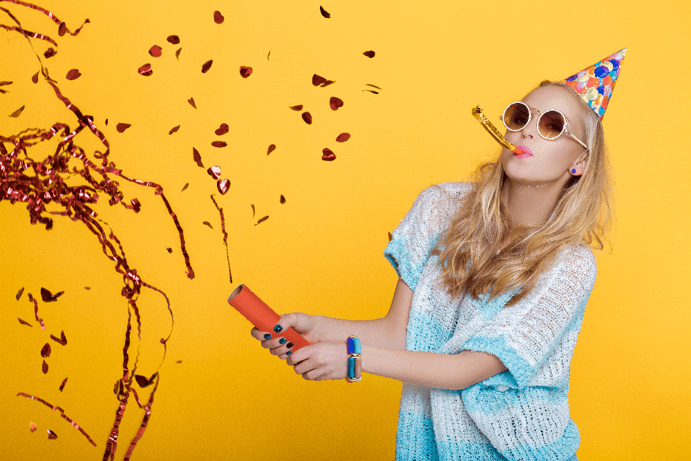 fête d'anniversaire explosion cottillon femme blonde fond jaune chapeau sur la tête langue belle mère