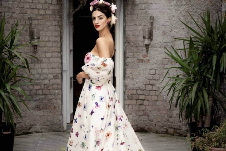 La robe est fluide et longue, avec une coupe élégante qui dégage les épaules, soulignant le cou et les clavicules. Les manches bouffantes tombent légèrement, ajoutant une touche romantique et vintage à la tenue. Le tissu est blanc cassé, parsemé de motifs floraux colorés dans des tons pastel et vifs, comme du rose, du violet, et du jaune femme porte une couronne de fleurs dans les cheveux, assortie aux motifs de sa robe, ce qui renforce l’ambiance florale et naturelle façade en briques pose avec élégance, légèrement de profil, tournée vers la caméra